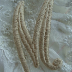 May include: A cream-colored embroidered letter M, stitched with multiple rows of thread, on a white lace background.