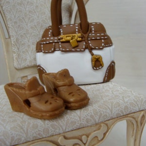 May include: A miniature brown and white handbag with a gold clasp and handle sits on a small chair. Two brown wedge sandals are in front of the bag. The chair has a cream-colored frame and a patterned seat.