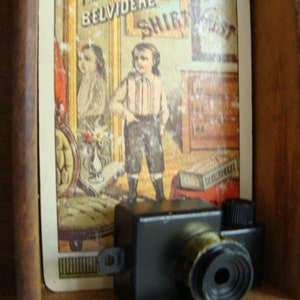 May include: A vintage toy camera with a black body and a silver lens. The camera is sitting on a wooden shelf with a vintage advertisement for "The Belvidere Shirt Waist" behind it.