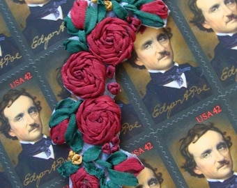 May include: A handmade fabric flower embellishment with red roses and green leaves. The embellishment is displayed on a background of postage stamps featuring the portrait of Edgar Allan Poe.
