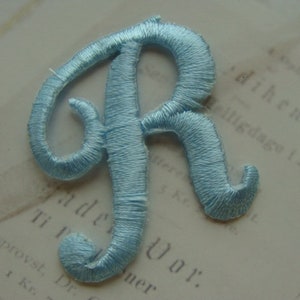 May include: A light blue embroidered letter R. The letter is stitched with a satin stitch and has a cursive style.