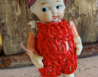 May include: A small porcelain doll with red yarn hair and a red crocheted dress. The doll has blue eyes and a painted smile.