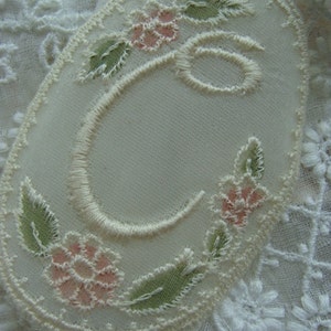 May include: Oval-shaped embroidered patch with the letter "C" in cream-colored thread. The patch is decorated with pink flowers and green leaves, outlined with a delicate cream-colored border. The background is a soft, off-white fabric.