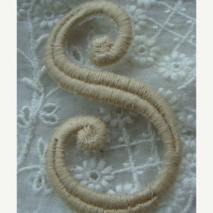 May include: A beige embroidered letter S, with a double-stitched outline, on a white lace background.
