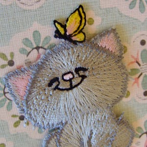 May include: Embroidered patch of a gray kitten with pink ears and a smiling face. A yellow and pink butterfly sits atop the kitten's head. The patch is on a light blue and floral patterned background.