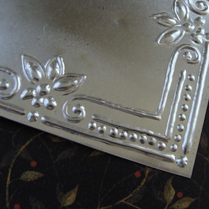 May include: A silver metal square with an embossed design around the border. The design features a repeating pattern of small circles and floral motifs.