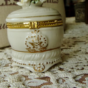 Puede incluir: Pequeña caja de baratijas de porcelana blanquecina con tapa floral y detalles dorados. La caja presenta una banda dorada decorativa alrededor del centro y un diseño dorado en la parte delantera. La base tiene un borde festoneado con detalles dorados.