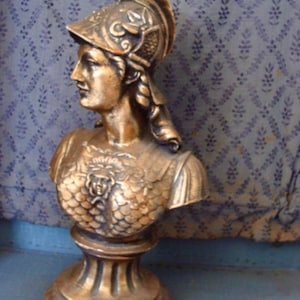 May include: A gold-colored bust sculpture of a woman wearing a helmet with a laurel wreath. The bust is on a pedestal with a round base.
