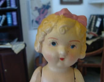 May include: A porcelain doll with blonde hair and a pink bow. The doll has blue eyes and a painted smile. The doll is wearing a white dress.