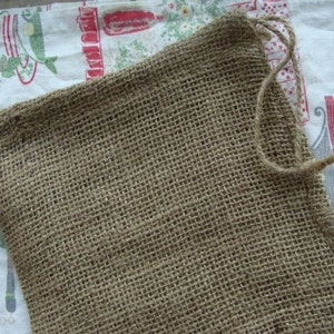May include: A brown burlap drawstring bag with a woven texture. The bag has a simple design with a drawstring closure made of twisted twine. The bag is empty and sits on a patterned fabric background.