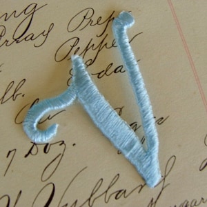 May include: A light blue felt letter V, embroidered with a satin stitch. The letter is on a white background with black handwriting.