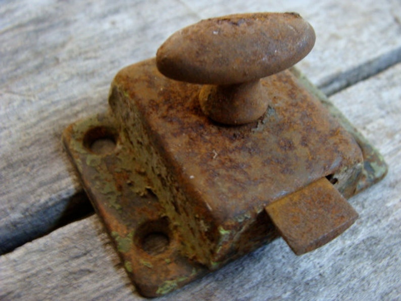 May include: A rusty metal latch with a rounded knob. The latch is green and brown with a rectangular shape. It has two holes on the side and a small metal tab on the bottom.