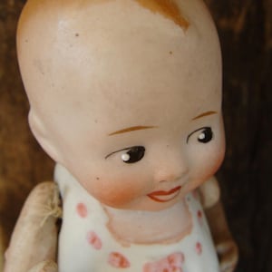 Puede incluir: Una muñeca de porcelana con un vestido blanco y detalles rosas. La muñeca tiene una cara pintada con cabello castaño y ojos azules.