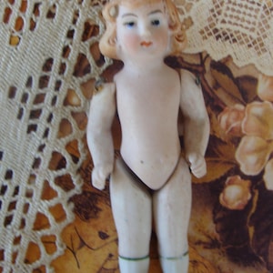 May include: A small antique porcelain doll with blonde curly hair, blue eyes, and rosy cheeks. The doll is wearing blue shoes and has green bands around its white socks. The doll's arms and legs are jointed.