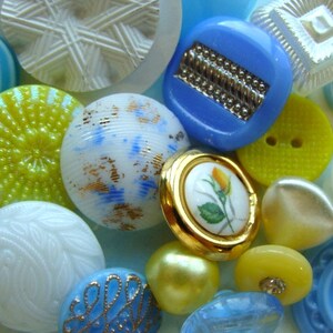 May include: Assortment of colorful buttons in various shapes, sizes, and patterns. The buttons are made of glass, metal, and plastic. Some buttons have a floral design, while others have geometric patterns. The buttons are a mix of blue, yellow, white, and gold.