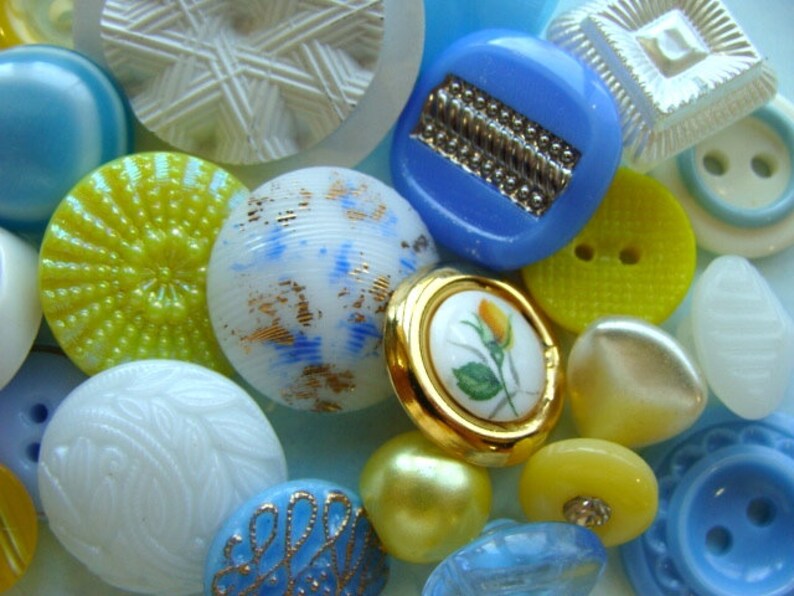 May include: Assortment of colorful buttons in various shapes, sizes, and patterns. The buttons are made of glass, metal, and plastic. Some buttons have a floral design, while others have geometric patterns. The buttons are a mix of blue, yellow, white, and gold.