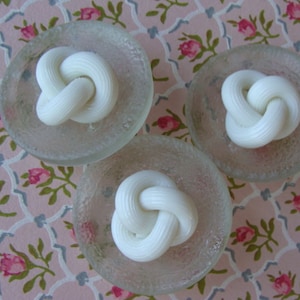 May include: Three white ceramic knots, each placed on a clear glass dish with a textured surface. The dishes are arranged on a pink and white floral patterned fabric.