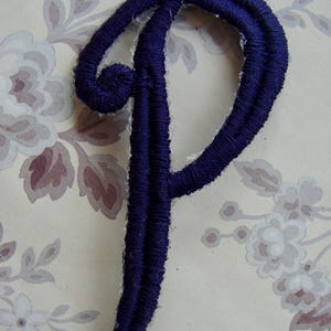 May include: A dark navy blue embroidered letter P patch featuring a cursive script font. The letter is constructed with dense thread stitching and has a visible white adhesive backing along the edges of the embroidered design.