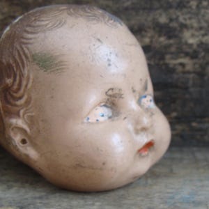 May include: A vintage porcelain doll head with painted blue eyes and a rosy blush. The doll's head is missing its body and has a chipped ear.