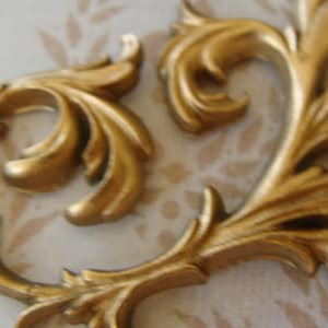 May include: Two ornate, gold-colored decorative flourishes with a detailed, swirling leaf design. The pieces have a metallic sheen and are set against a patterned background, suggesting a vintage or antique aesthetic.