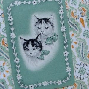 May include: A vintage greeting card with a green background and a white floral border. Two black and white kittens are featured on the card, one with a white bow around its neck.