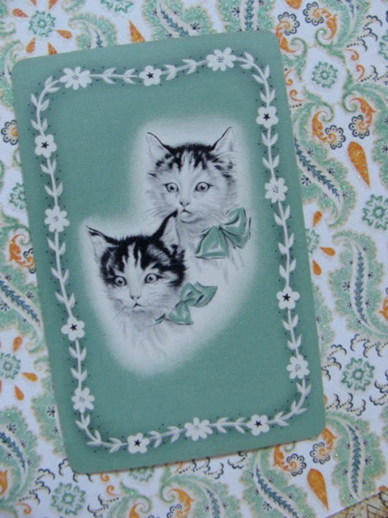 May include: A vintage greeting card with a green background and a white floral border. Two black and white kittens are featured on the card, one with a white bow around its neck.