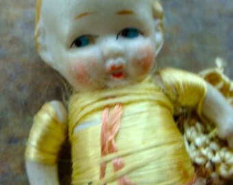 May include: A porcelain doll with a light brown wig, blue eyes, and a pink and white dress. The doll is wearing a yellow and white fabric wrap around its torso.