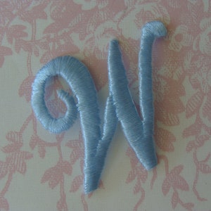 May include: A light blue embroidered letter W. The letter is on a white background with a pink floral pattern.