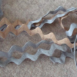 May include: Close-up of wavy ribbon in shades of gray and tan. The ribbon has a zigzag edge and is arranged in a layered pattern against a textured, neutral background. This decorative ribbon is suitable for crafts and sewing projects.