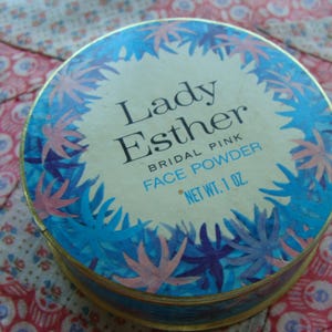 May include: A round, blue and pink compact with a white label that reads "Lady Esther Bridal Pink Face Powder Net Wt. 1 oz."