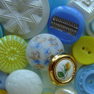 May include: Assortment of colorful buttons in various shapes and sizes, including a blue button with a silver design, a yellow button with a raised center, and a white button with a swirling pattern.