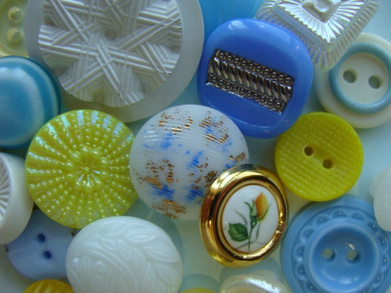 May include: Assortment of colorful buttons in various shapes and sizes, including a blue button with a silver design, a yellow button with a raised center, and a white button with a swirling pattern.