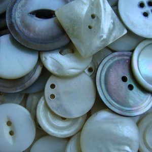 May include: A collection of white and gray pearl buttons of various sizes and shapes. Some buttons have two holes, while others have four holes. The buttons are arranged in a random pattern.