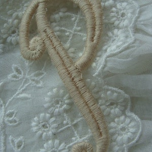 May include: A beige embroidered letter P, with a double-stitched outline, on a white lace background.