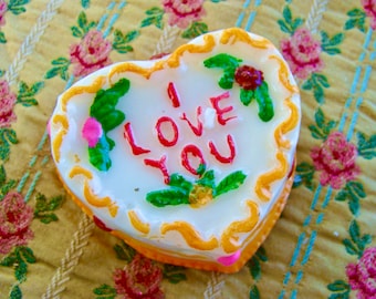 May include: A white heart-shaped cake with yellow icing and a red and white "I LOVE YOU" message. The cake is decorated with green leaves and pink flowers.