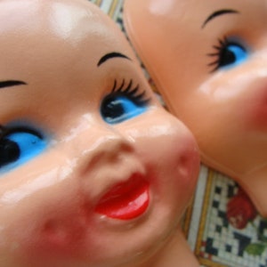 May include: Two porcelain dolls with painted blue eyes and red lips. The dolls have a white and pink complexion and are wearing no clothing.