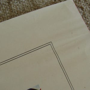 May include: A corner of a cream-colored paper with a double black border. The paper has slight imperfections and is set against a textured, light brown surface.