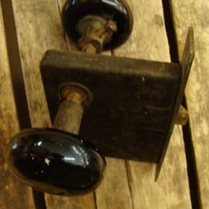 May include: A black door knob with a round, black porcelain handle and a metal base. The knob is attached to a wooden door.
