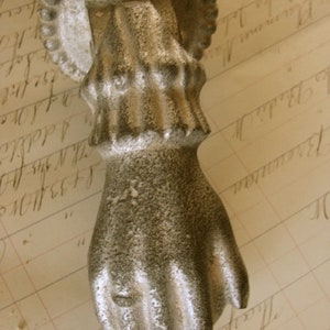 May include: A vintage-style door knocker in the shape of a hand, cast in a silver-toned metal. The hand is detailed with textured surfaces and a ring on the ring finger. The knocker is mounted on a circular backplate with a beaded edge.