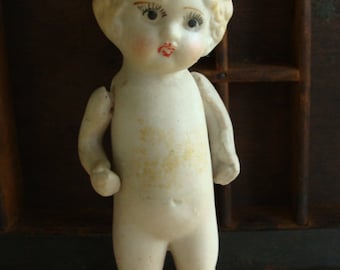 May include: A porcelain doll with a white dress and a red headband. The doll has painted eyes, a red mouth, and a rosy complexion.