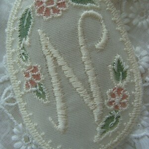 May include: A white oval embroidered with a large capital "N" in white thread, surrounded by pink and green floral embroidery.