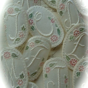 May include: A collection of off-white fabric appliques with embroidered letters and floral designs. The letters are in a script font and the flowers are pink and green.