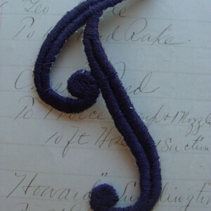 May include: A dark purple fabric letter J with a double stitched outline. The letter is on a white piece of paper with handwritten text.