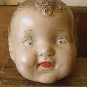 May include: A vintage porcelain doll head with painted blue eyes and a painted smile. The doll's hair is sculpted and painted to look like a curly hairstyle.