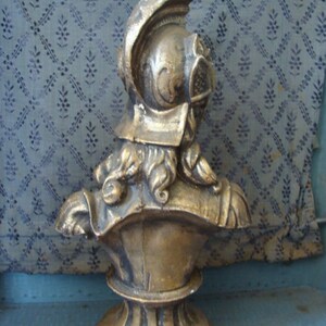May include: A gold-colored bust sculpture of a person wearing a helmet. The bust is on a pedestal with a round base.