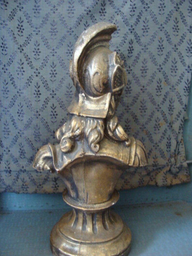 May include: A gold-colored bust sculpture of a person wearing a helmet. The bust is on a pedestal with a round base.