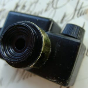 May include: A small black plastic toy camera featuring a circular lens barrel with concentric rings and a textured gold-colored metal ring at the base. The camera body has a rectangular shape with a prominent black dial positioned on the top right corner.