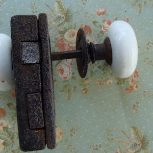 May include: Antique door hardware set featuring a weathered, dark brown metal latch and two white oval porcelain door knobs. The hardware is designed for a door and is set against a floral patterned background.