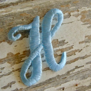 May include: A light blue embroidered letter H. The letter is stitched with a cursive font and is on a white wooden background.