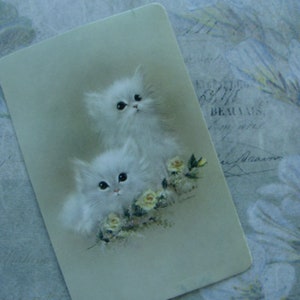 Op de afbeelding: Een vintage kaart met twee witte kittens met blauwe ogen, omringd door witte bloemen. De kittens knuffelen elkaar en hebben een zacht, pluche uiterlijk.